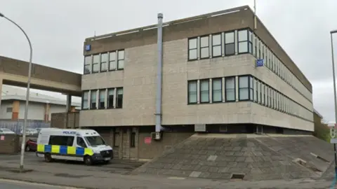 Google Street view image of Washington Police station, on the corner of two streets with police van parked in front. It's a large, brutalist, grey building.