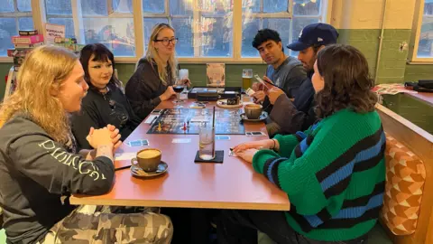 Six young people sitting at a cafe table playing Cluedo. as well as the game pieces, there are various drinks on the table. In the background is a large window