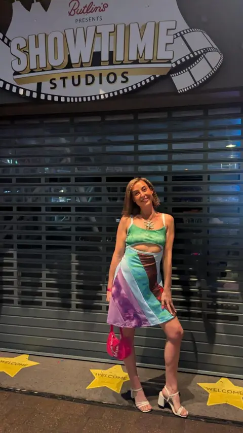 Charlotte Morjaria A woman wearing a multicoloured, striped skirt and green crop top, with white heels and a pink handbag. She's stood smiling, in front of a shutter, with yellow stars on the ground and a sign above her which reads: Butlin's presents showtime studios".