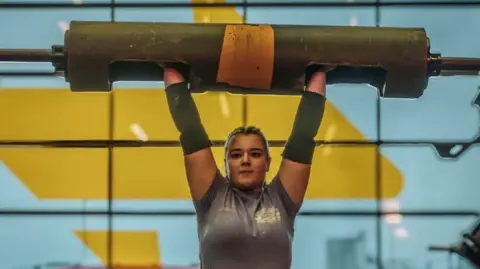 A close-up shot of teenager Lauri Siegal, holding a large weight overhead. She is wearing a grey T-shirt and has a determined expression. A wall of windows with yellow graphics is behind her.