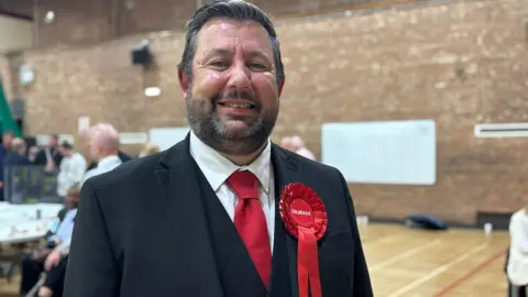 Claire Hamilton/BBC Anthony Burns, in his late 40s or early 50s, with black and grey hair styled into a quiff and a light beard, smiles at the camera while wearing a black suit, white shirt and red tie. He has a Labour rosette pinned to his lapel.