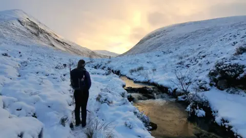 Rory Dracup A woman stands with her back to the camera in a snowy landscape. a river runs down past her left hand side. In front of her is a dramatic mountain pass.