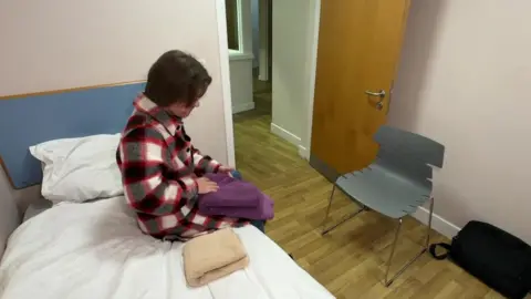 Adam is sat on a bed in a bedroom at the shelter folding towels