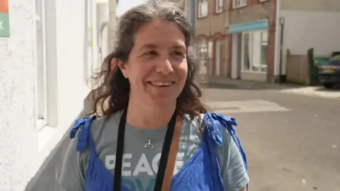 A woman wearing a grey top and blue dungarees over the top and a silver necklace. She is smiling at the camera. She has brown hair which is tied back. Behind her is a street.