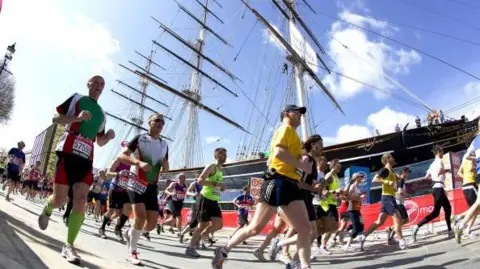 Getty Runners take part in the London Marathon in Greenwich, passing a historic sailing ship while spectators line the roadside.
