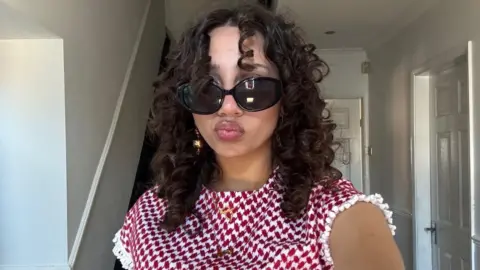 Mariam Azam Mariam smiles at the camera and is wearing sunglasses with a white and red top. She has curly brown hair 