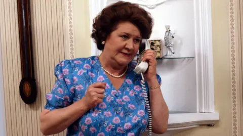 Patricia Routledge is seen playing Hyacinth Bucket in Keeping Up Appearances. She is wearing a blue floral dress and pearl necklace and holding a corded telephone, with decorative shelving and patterned wallpaper visible behind.