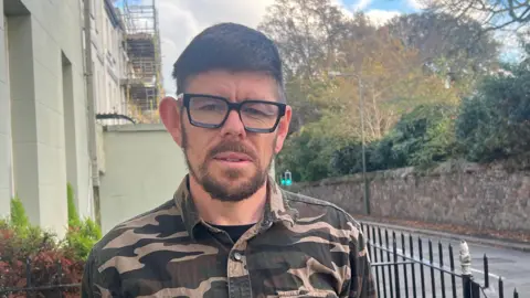 Daniel Parkes looks at the camera. He's wearing a camo shirt, with large black framed glasses. He has a short brown beard and dark hair. He is standing in the front garden of his home, with the roadside visible close by. 