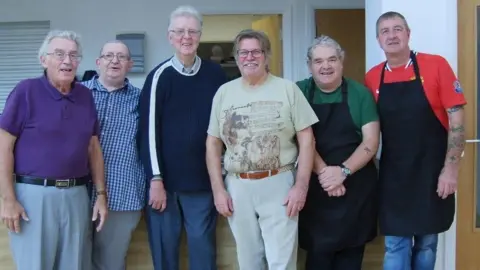 Six men are standing together and smiling next to the charity's kitchen. Two of them are wearing dark-coloured aprons. 