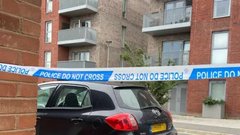 A black car parked by a set of flats. There is police tape in front of the car and flats and it reads Police do not cross. 