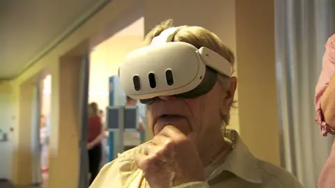 A man with grey hair is wearing a white virtual reality set. His hand is on his chin, reflecting he ponders what he sees and his mouth is slightly ajar