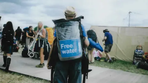 WaterAid A film photo showing a WaterAid volunteer from the back, wearing a large bag which says 'free water' on it. In front of him there are people carrying tents and luggage towards the campsite.