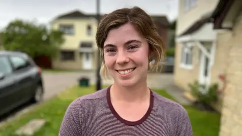 Women smiling at camera with dark brown hair. She's wearing a stripey top and stood outside a house in the garden.