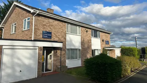 A semi-detached two-storey house with white panelling under windows and a drive in front of a white garage door. A small front lawn has bushes in front of it. A dark blue sign above the door says Crime Scene House. The sky is blue.