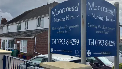 A two-storey care home which is inside what appears to have once been houses. The sign for the care home is in the forefront and reads: "Putting the older person's interest first." It also has the care home's name and contact details on it.