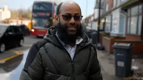 Abu Shah is bald, has a big black beard and is wearing black sunglasses and a black puffer-style coat