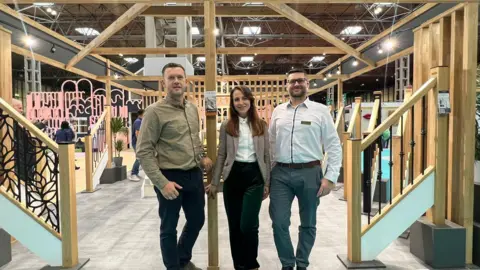 Lukasz Garbacik Lukasz Garbacik poses for a photo with his wife Wioletta and brother Tomasz. Lukasz is wearing a beige-coloured shirt with blue jeans and has short, fair hair. Wioletta is wearing a cream blazer with dark trousers and a white shirt. She has long brown hair. Tomasz, in grey trousers and a white shirt, has short black hair and is wearing glasses.