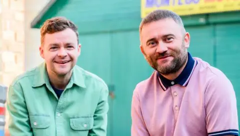 River City characters Angus Lindsay (played by Scott Fletcher) and Bob O'Hara (played by Stephen Purdon) smile at the camera wearing casual clothes outside of a garage