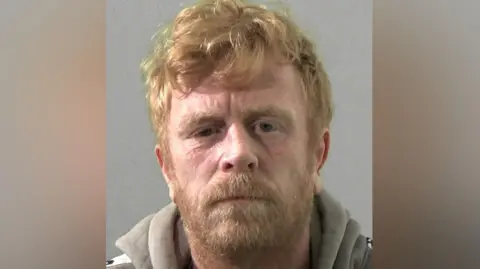 Mugshot of Black. He has scruffy ginger hair and a beard and is wearing a light grey hoody