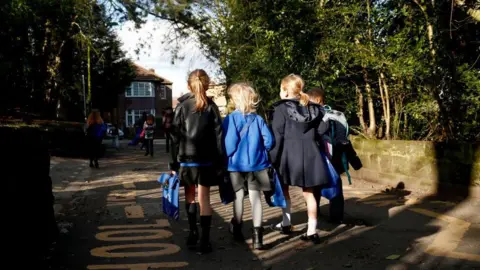 Three small girls and a boy pictured from behind walking along a pavement on which the word school is painted in large yellow capitals. All wear blue and black school uniform. It is a sunny day.