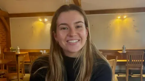 Kaitlin Lawrence has long blond hair and is wearing a black jumper. She is smiling and sitting in a cafe.