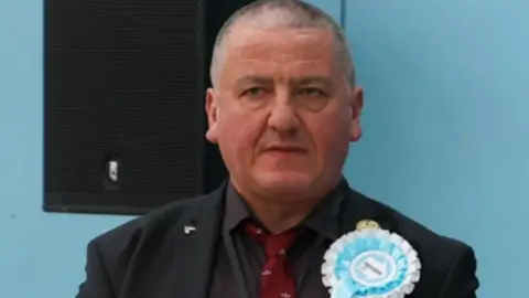 Andrew Harrison is wearing a black suit with a dark red tie with a light blue Reform rosette. He has close cropped grey hair.