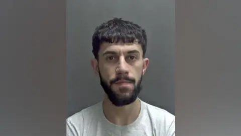 Safa Lawan is looking at the camera in a police custody image. He has dark, short hair and a dark beard and moustache. He is wearing a grey T-shirt