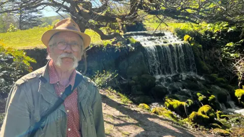 Doug Chalk posing next to the rocky waterfall at Littlebredy. It's a bright sunny day and he is wearing a mustard colour trilby hat, a plum collar shirt with small white spots and a khaki green jacket. He has a large grey moustache and round glasses.