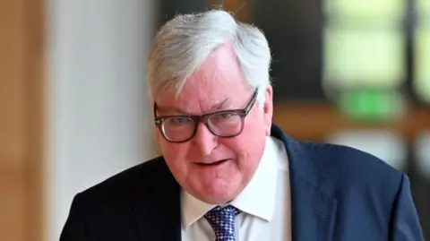 A bespectacled man with white hair in a dark blue suit