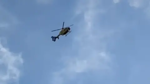 Sophie May A yellow air ambulance helicopter photographed from afar - background is blue sky with clouds.