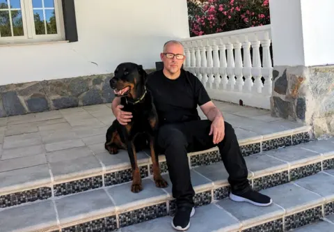 Ryan O'Meara Ryan O'Meara sitting on a step outside a house with his dog. He wears a dark t-shirt and jeans and black trainers with white soles. He is wearing glasses. His dog is large, brown and black and with big teeth. 