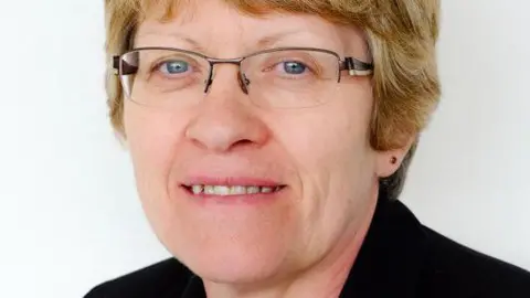 A head shot image of a 67-year-old woman smiling. She is wearing glasses and has blonde hair. The background is white. 