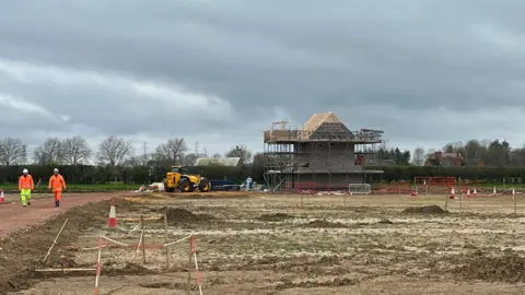 The construction of a building is happening in a muddy field. The house has its walls built but the roof has exposed rafters and the whole building has scaffolding around it. There is a large plant vehicle next to the building and the area is protected by red and white barriers and cones. To the left, there are two construction workers, wearing high-vis trousers and jackets and hard hats, are walking towards the camera. 
