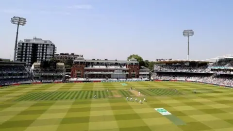  The arc of a stand, and two floodlights, behind a green cricket field. A bowler is at the far crease delivering a ball to a batsman. A wicketkeeper and four slips are crouched behind the batsman. Fielders can also be seen at cover point, mid-off, mid-on and mid-wicket. One umpire stands next to the bowler and another umpire stands at square leg.