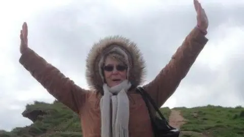 Supplied A woman in her seventies is pictured with her arms in the air. She is wearing a brown coat with a fur-lined hood pulled up over her head and a grey scarf. She is standing on a hill with a path going uphill behind her.