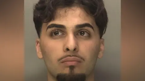 Deara Sadiq, pictured in a police mugshot. He is a man with dark brown hair and a well-groomed goatie beard and well as a thin moustache.