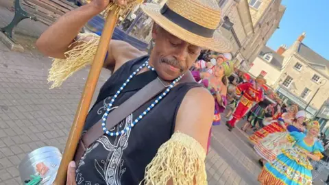Stroud Valleys Artspace Image of a man wearing a straw boater and a black vest top. He is holding a long pole. On his arms he is wearing cuffs of straw and around his neck he has a string of blue beads. In the background can be seen a a group of dancers wearing brightly coloured long ethnic print dresses.