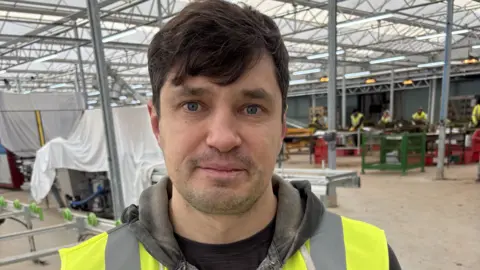 Alex Sivko, is a clean shaven, dark-haired young man in a grey hoodie and yellow tabard. He is standing in the middle of the shot in a metal framed industrial building. 