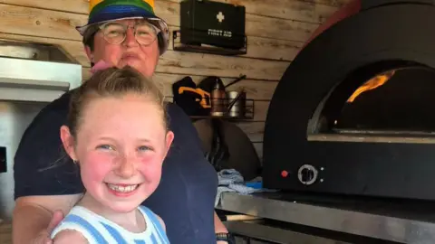 Woo_reviews Woo, smiling at the camera, has her hair tied back and is wearing a blue and white dress. A woman is standing behind her, wearing a blue top, a hat and glasses. A pizza oven is on the right.