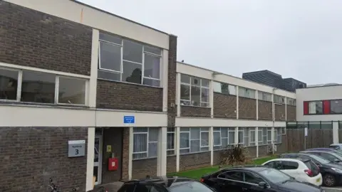 A dark brown bricked building goes across the image. The two-storey building has multiple windows and an entrance on the left. Cars are parked outside and a stretch of grass goes alongside the building.