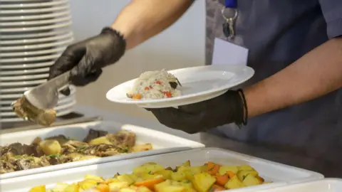 Gambar getty seseorang yang menyajikan makanan panas ke atas piring di atas meja yang mengenakan celemek dan sarung tangan