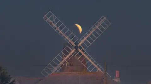 A moon is seen between blades of a windmill.