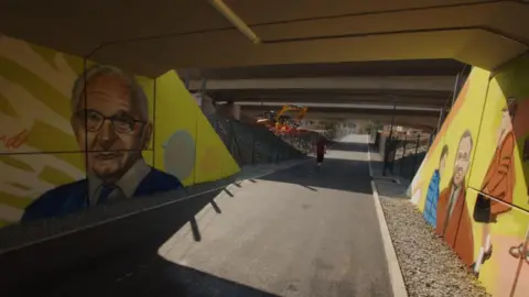 BBC View of Nicholas Winton thoroughfare  with murals connected  walls eithers side, including 1  representation  of Nicholas Winton