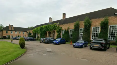 Exterior shot of an ivy-clad building with large Georgian-style windows. There are a number of cars parked in front of the building, along with a lawned area and shrubs.