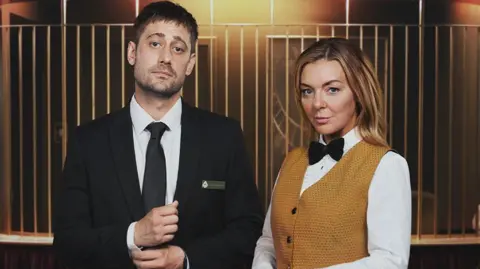 Left: Matty (played by actor Michael Socha) dressed as a croupier wearing a black suit in TV drama The Cage alongside his co-star Leanne (Sheridan Smith) wearing a mustard coloured waistcoat and white shirt. The scene is set in a casino.