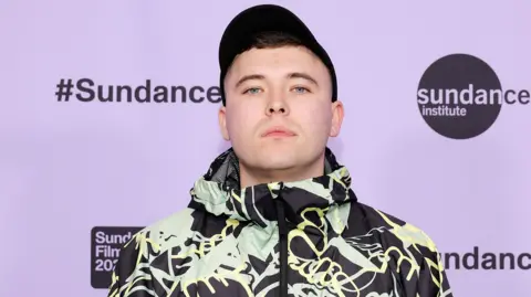 Getty Images A dark haired young man with short brown hair and blue eyes, wearing a black cap and a patterned coat. The backdrop behind him is lilac and says Sundance.
