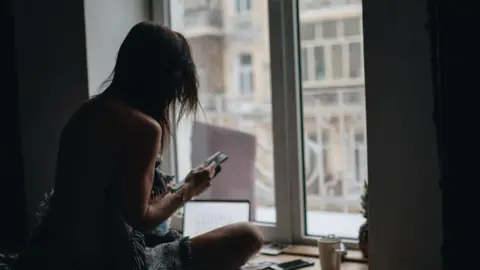 A woman in silhouette sitting in front of a window looking at her phone