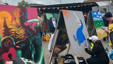 Upfest This picture has three painters designing their pieces in a courtyard. They are designing on large boards. One painting has a man wearing a mask to breath with a purple/red sunset behind him. The painting in the middle has a blue pelican bird looking at the sun. The painting at the far end appears to be a beach and the ocean on a sunny day.