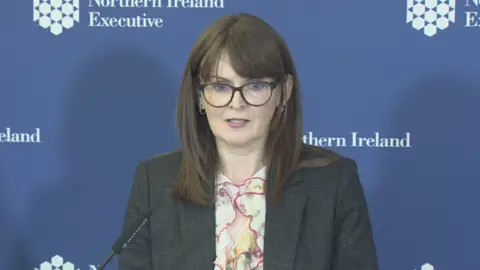 The finance minister wearing a dark grey suit and floral shirt. She stands infront of a blue board with the Northern Ireland executive logo. 