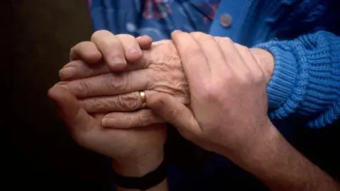 Two hands clasped around the hands of an elderly woman wearing a wedding ring and a blue cardigan.